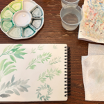 Watercolor supplies, including an open sketchbook, porcelain palette, cups of water, and blotting paper. The sketchbook shows bunches of green leaves, painted in watercolor.